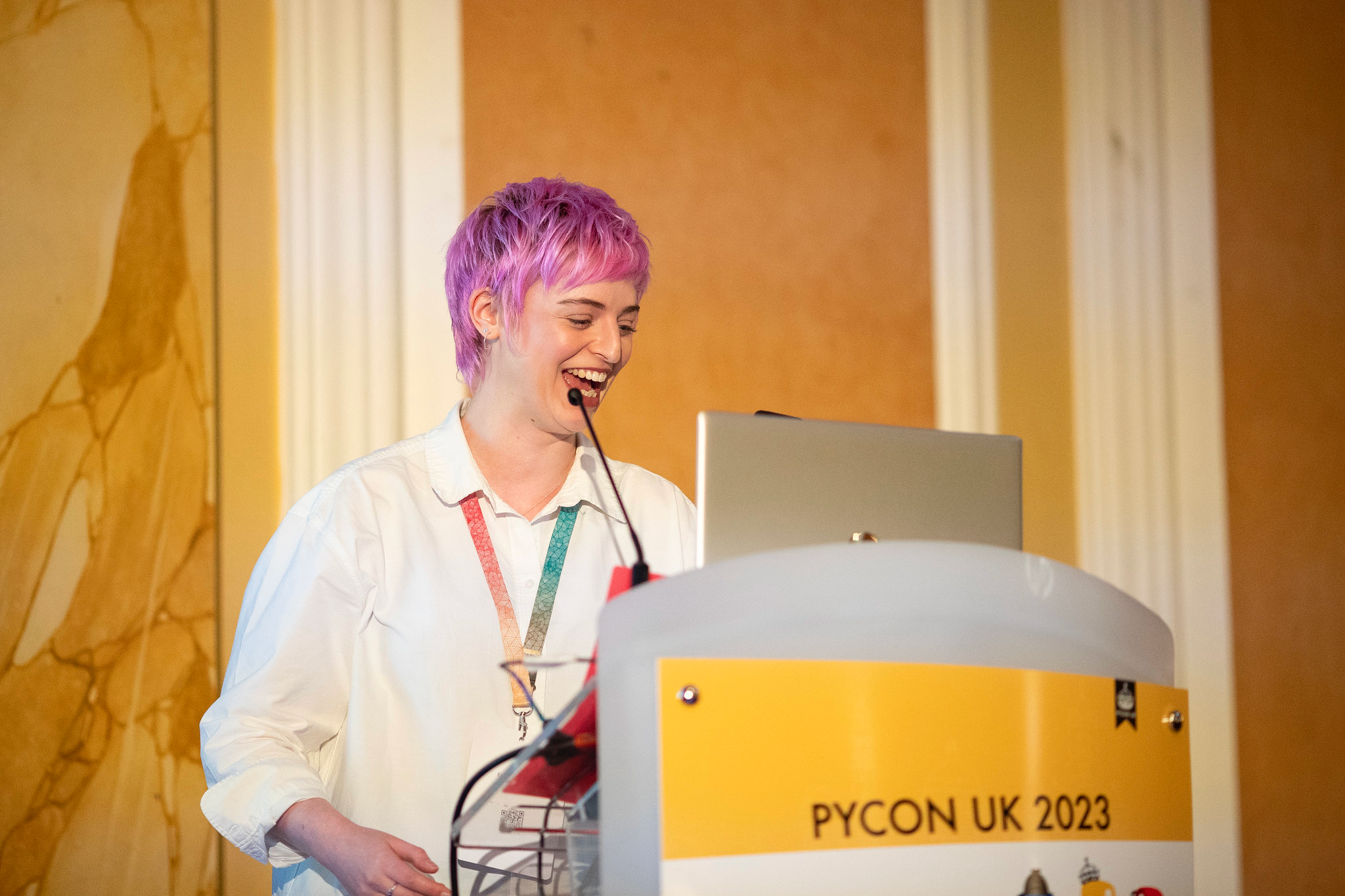 Emily, a white woman in her thirties with pink hair, laughing as she speaks from a PyCon UK 2023 podium